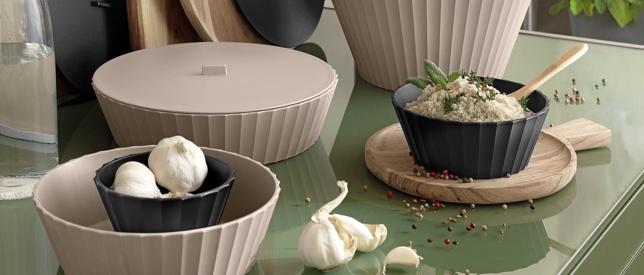 Contenitori Hera Blim Plus su tavolo verde con aglio e verdure in ciotole Carbon Black e Brown Sugar